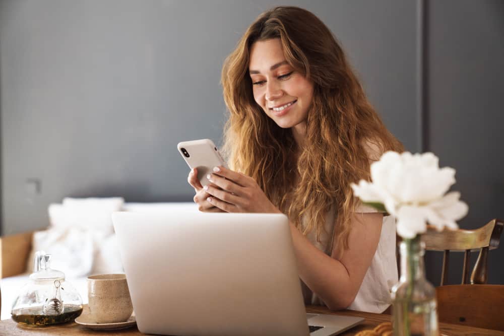 Woman sits at table with laptop, smiling at mobile phone