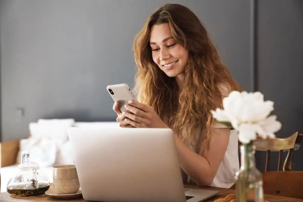 Woman sits at table with laptop, smiling at mobile phone